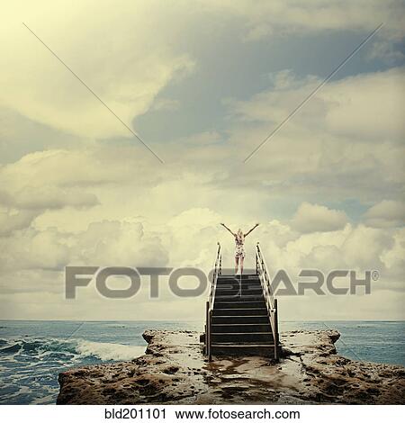 Stock Image - Caucasian woman on stairs on rocks next to ocean. Fotosearch