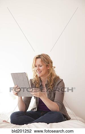Stock Image - Caucasian woman sitting on bed using digital tablet. Fotosearch