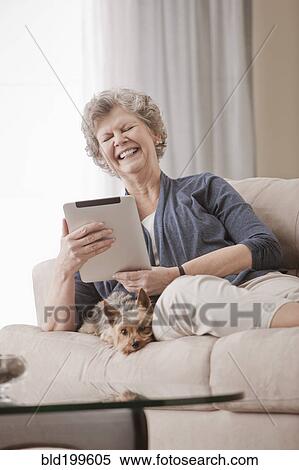 Stock Photography - Caucasian woman sitting on sofa using digital tablet with dog. Fotosearch