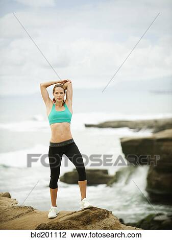 Stock Image - Caucasian woman stretching on rocks near ocean. Fotosearch