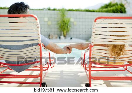 Stock Image - Couple holding hands in lounge chair near swimming pool. Fotosearch