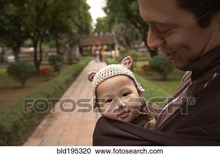 Stock Image - Father carrying baby in sling in park. Fotosearch