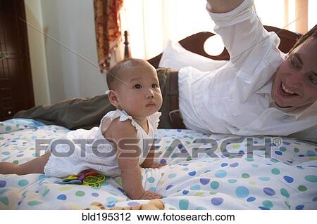 Stock Image - Father playing with baby girl on bed. Fotosearch