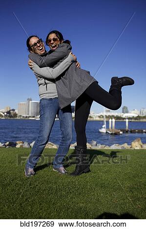 Stock Photo - Grinning mixed race sisters hugging outdoors. Fotosearch