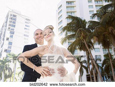 Stock Image - Hispanic bride in groom hugging outdoors. Fotosearch