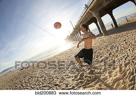 Stock Image - Hispanic man playing with soccer ball on beach. Fotosearch