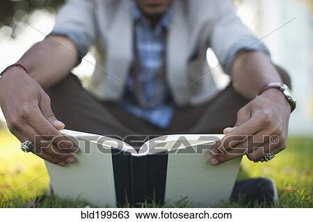 Stock Image - Hispanic man reading book in grass. Fotosearch