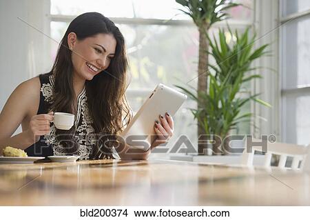 Picture - Hispanic woman drinking coffee and using digital tablet. Fotosearch