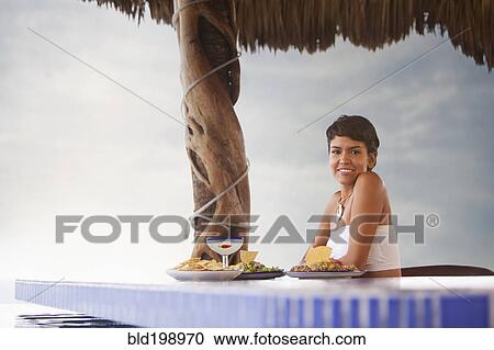 Stock Image - Hispanic woman having dinner at poolside. Fotosearch