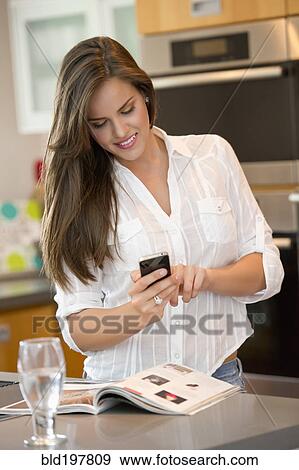 Stock Photo - Hispanic woman using cell phone in kitchen. Fotosearch