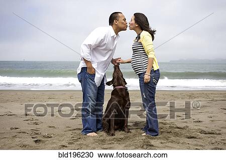Stock Image - Husband kissing pregnant wife on beach while dog watches. Fotosearch