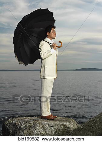 Stock Image - Japanese businessman holding umbrella near ocean. Fotosearch