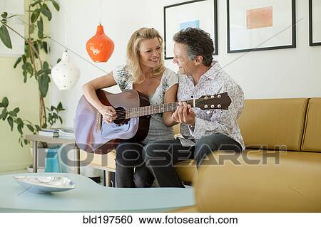 Stock Image - Man teaching wife to play guitar in living room. Fotosearch