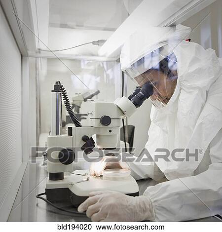 Stock Image - Middle Eastern scientist in clean suit using microscope. Fotosearch