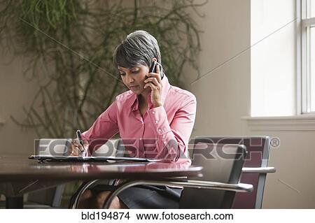 Picture - Mixed race businesswoman writing and using cell phone. Fotosearch