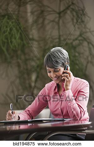 Stock Photography - Mixed race businesswoman writing and using cell phone. Fotosearch