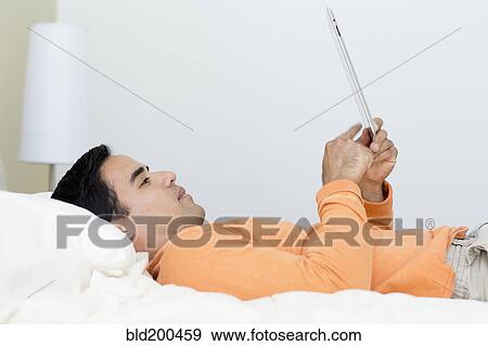 Stock Photo - Mixed race man laying in bed using digital tablet. Fotosearch