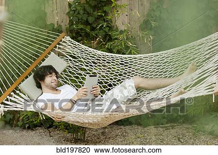 Stock Image - Mixed race man laying in hammock using digital tablet. Fotosearch