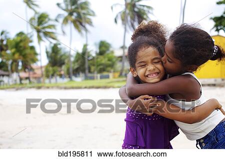 Stock Image - Mixed race sisters hugging on beach. Fotosearch