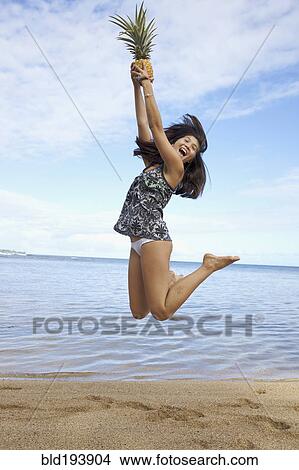 Picture - Mixed race teenager jumping and holding pineapple on beach. Fotosearch