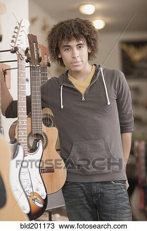 Stock Image - Mixed race teenager standing with guitars in music store. Fotosearch