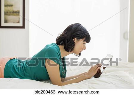 Stock Image - Mixed race woman laying on bed using digital tablet. Fotosearch