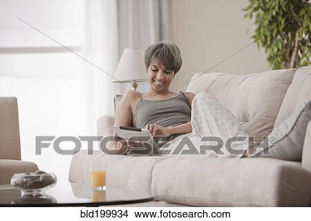 Picture - Mixed race woman on sofa using digital tablet. Fotosearch