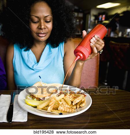 Stock Photo - Mixed race woman squeezing ketchup on french fries in diner booth. Fotosearch