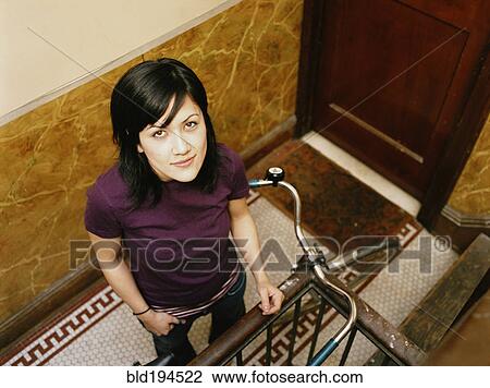 Stock Image - Mixed race woman standing with bicycle in apartment hallway. Fotosearch