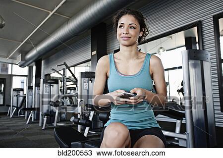 Stock Image - Mixed race woman text messaging on cell phone in health club. Fotosearch
