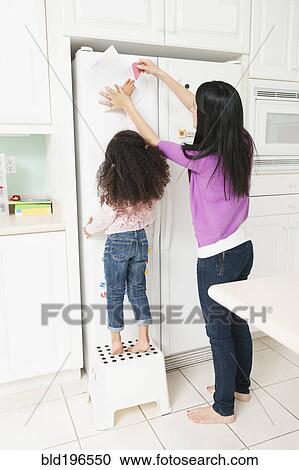 Stock Image - Mother putting daughter's drawing on refrigerator. Fotosearch
