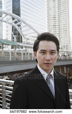 Stock Image - Serious Asian businessman standing outdoors. Fotosearch