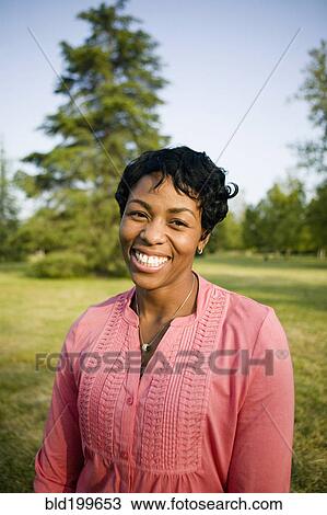 Stock Image - Smiling Black woman standing in park. Fotosearch