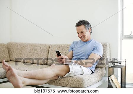 Stock Photography - Smiling man text messaging on cell phone in living room. Fotosearch
