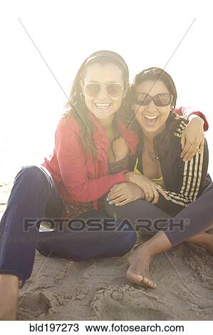 Stock Image - Smiling mixed race sisters sitting on beach. Fotosearch