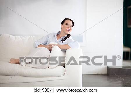 Stock Image - Smiling woman sitting on sofa drinking coffee. Fotosearch