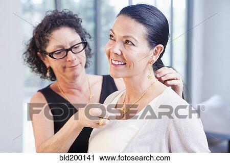Stock Image - Woman helping customer put on necklace in jewelry store. Fotosearch