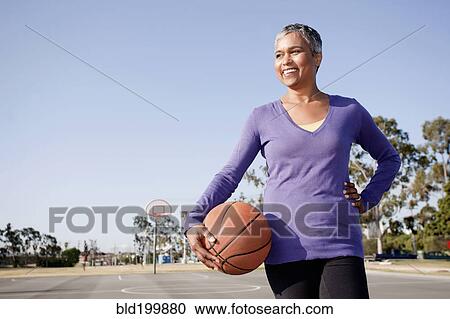 Stock Image - Woman holding basketball on basketball court. Fotosearch