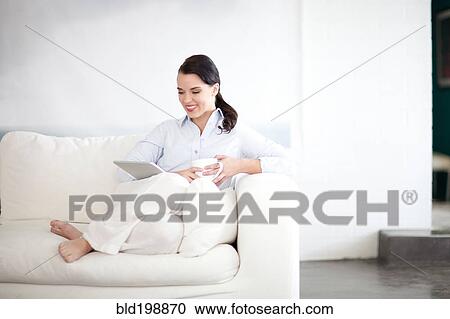 Stock Image - Woman sitting on sofa using digital tablet. Fotosearch