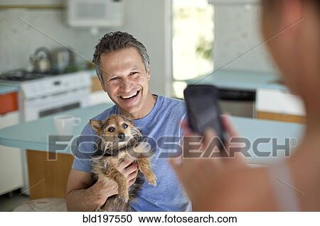 Stock Image - Woman taking picture of husband holding dog in kitchen. Fotosearch
