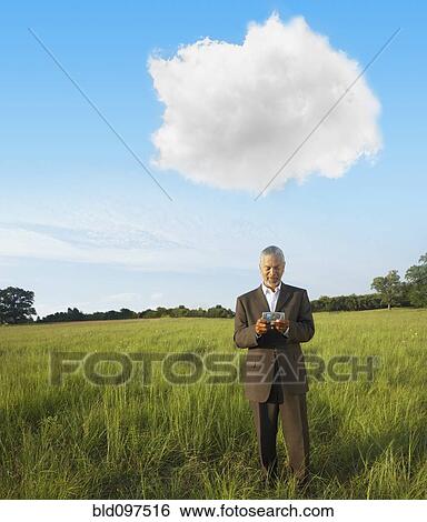 Stock Photograph - Black businessman standing in field text messaging on cell phone. Fotosearch