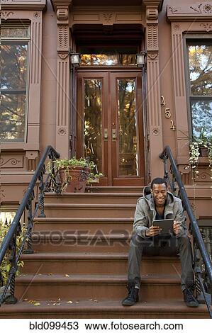 Stock Image - Black man sitting on front stoop using digital tablet. Fotosearch