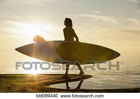 Stock Image - Caucasian man carrying paddle board on beach. Fotosearch