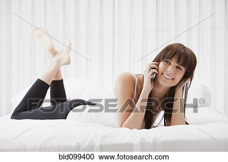 Stock Image - Caucasian woman laying on bed text messaging on cell phone. Fotosearch