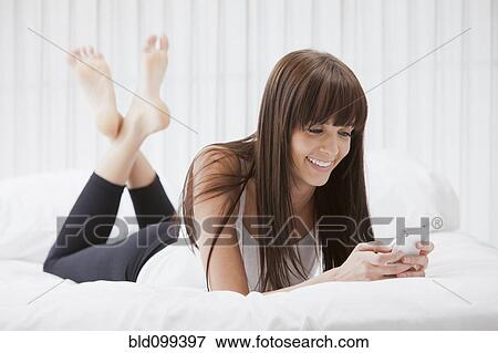 Stock Photo - Caucasian woman laying on bed text messaging on cell phone. Fotosearch
