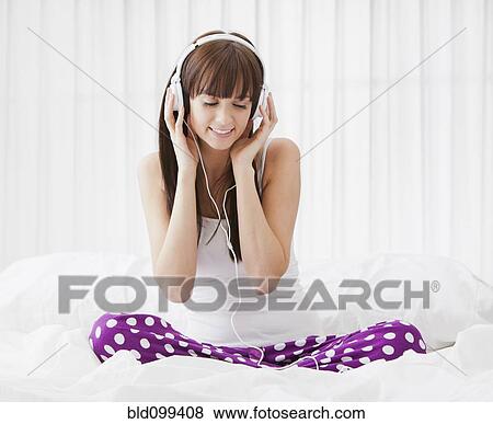 Stock Photo - Caucasian woman sitting on bed listening to music on headphones. Fotosearch