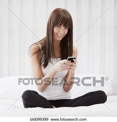 Caucasian woman sitting on bed text messaging on cell phone View Large Photo Image Stock Photo - Caucasian woman sitting on bed text messaging on cell phone. Fotosearch