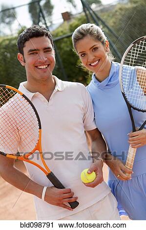Stock Image - Hispanic couple standing on tennis court with racquet and ball. Fotosearch