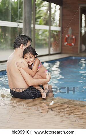 Stock Image - Hispanic father and daughter sitting on edge of swimming pool. Fotosearch