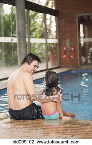 Stock Image - Hispanic father and daughter sitting on edge of swimming pool. Fotosearch